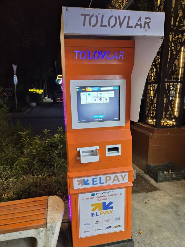 Оранжевый платёжный терминал ELPAY «To'lovlar» вечером у скамейки: оплата мобильной связи (Uzmobile, Beeline, Mobiuz, Ucell), интернета Perfectum, коммунальных услуг, такси, пополнение карт Uzcard и Humo; наклейка «Paynet — с нами платить быстро и удобно!»