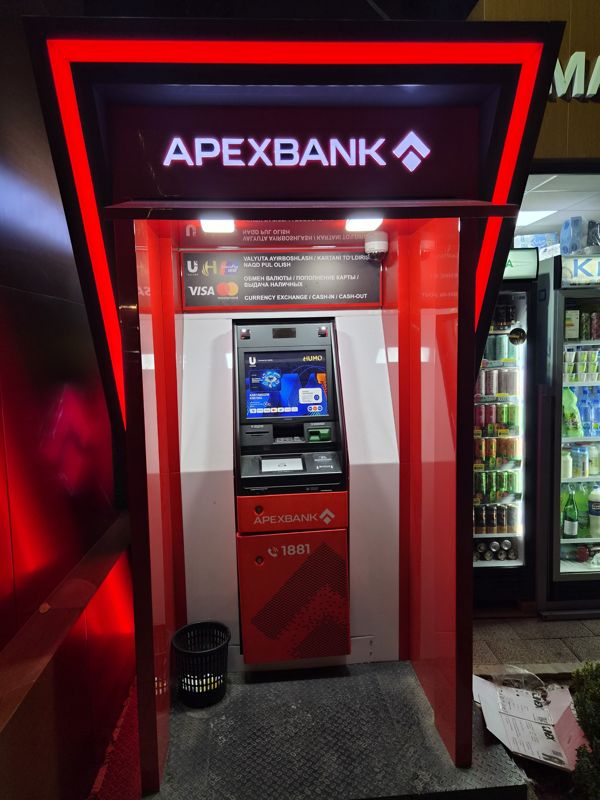 Банкомат Апексбанка в красной будке с подсветкой вечером, у витрины магазина: обмен валюты, пополнение карты, выдача наличных; карты Uzcard, Humo, Visa, Mastercard