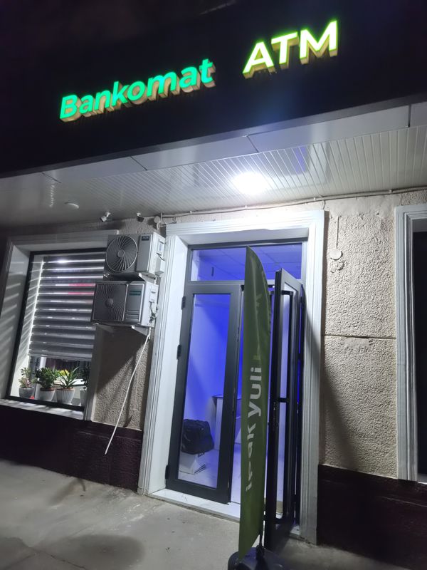 Светящаяся вывеска «Bankomat ATM» над входом в помещение с банкоматами; у двери флаг Ipak Yuli Bank