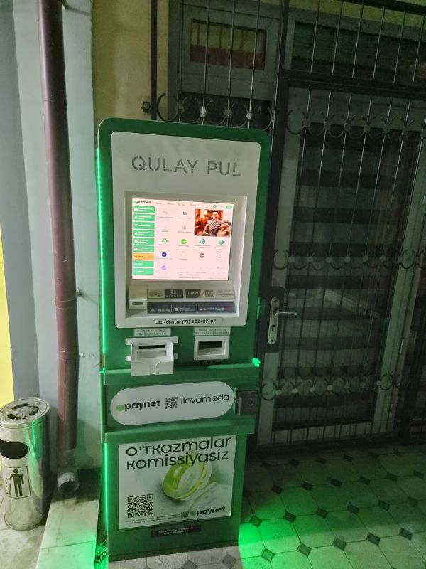 Платёжный терминал Paynet «Qulay pul» у решётчатой двери: экран с услугами, приём наличных, лоток чека; рядом урна
