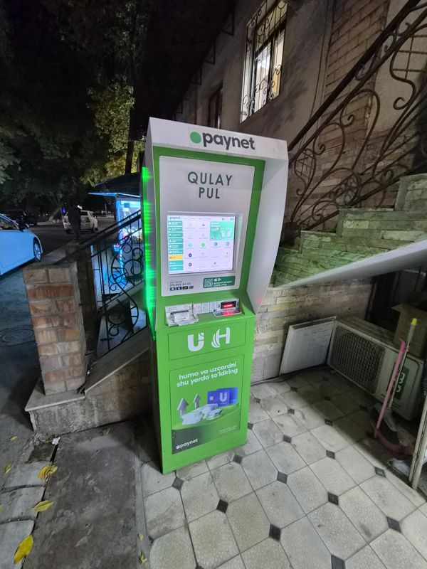 Платёжный терминал Paynet «Qulay pul» на тротуаре у лестницы старого здания: экран с услугами, лоток чека, купюроприёмник