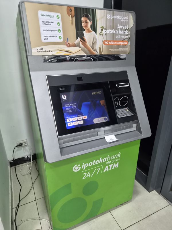 Банкомат Ипотека-банка (OTP Group, 24/7 ATM, NCR) в углу помещения: экран приветствия Humo / Uzcard, сверху реклама микрозайма до 100 млн сум