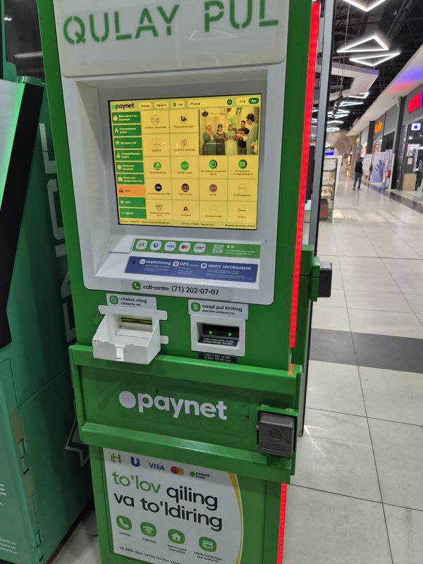 Платёжный терминал Paynet «Qulay pul» в проходе торгового центра: экран с услугами, лоток чека, купюроприёмник; слева край соседнего банкомата