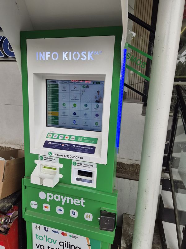 Платёжный терминал Paynet «Info Kiosk 24/7» под навесом у входа магазина: сенсорный экран, выдача чека и купюроприёмник