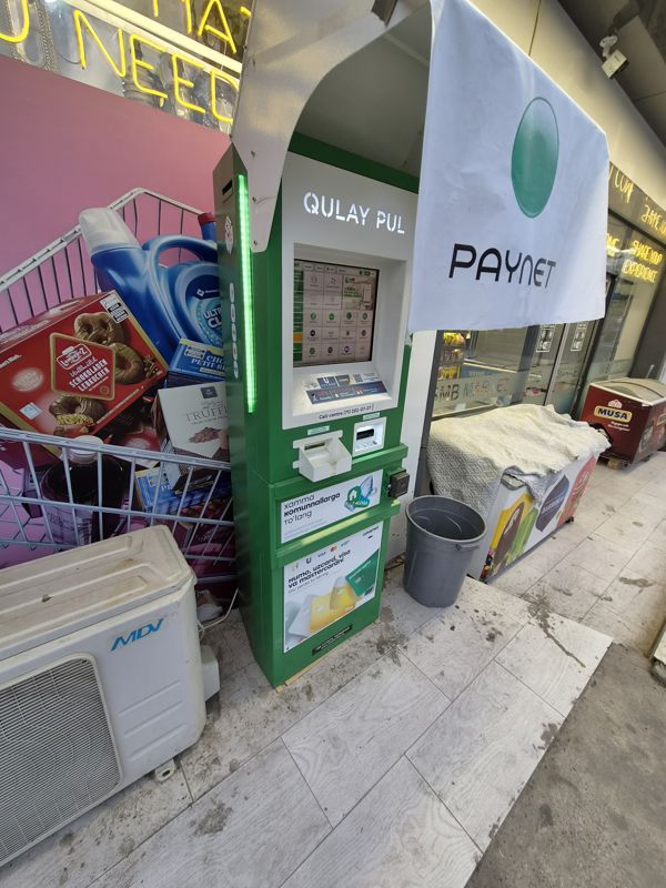 Платёжный терминал Paynet «Qulay Pul» с зелёной подсветкой у входа в BMB Market: оплата коммунальных услуг и пополнение карт Humo, Uzcard, Visa, Mastercard; сверху полотнище Paynet