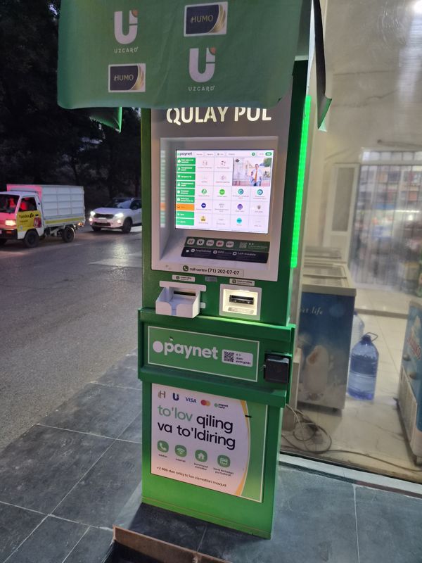 Вечером: платёжный терминал Paynet «Qulay Pul» у стеклянной витрины магазина на улице; сверху полотнище с логотипами Uzcard и Humo, на дороге грузовичок Yandex Go и автомобиль