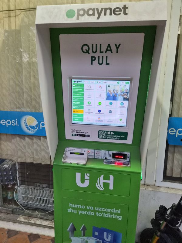 Платёжный терминал Paynet «Qulay Pul» внутри магазина у входа: экран с услугами, выдача чека, купюроприёмник; на стекле наклейки Pepsi