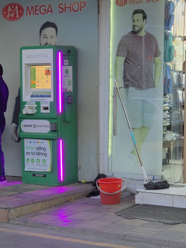 Вечером: платёжный терминал Paynet «Qulay Pul» у входа в магазин Mega Shop на тротуаре, по бокам фиолетовая подсветка; наклейка Paynet с QR-кодом и плакат «to'lov qiling va to'ldiring»