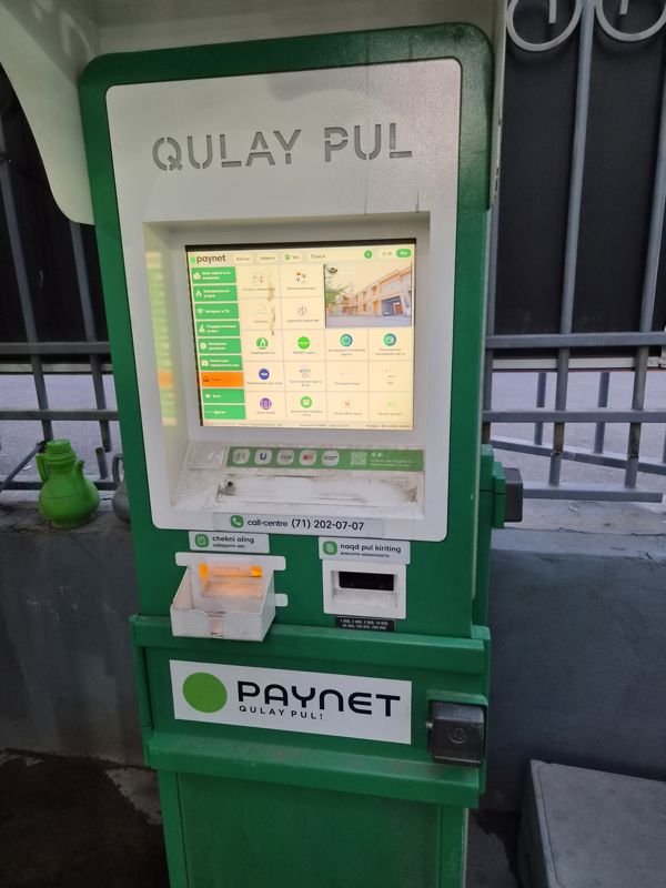 Платёжный терминал Paynet «Qulay Pul» у металлической ограды: экран с услугами, выдача чека, купюроприёмник, наклейка «PAYNET Qulay pul!»