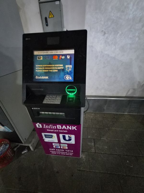 Банкомат InfinBANK у белой стены с электрощитом: экран «Iltimos kartangizni kiriting», бесконтактный считыватель, купюроприёмник, фиолетовая панель с Humo, Uzcard, Visa, «Мир»