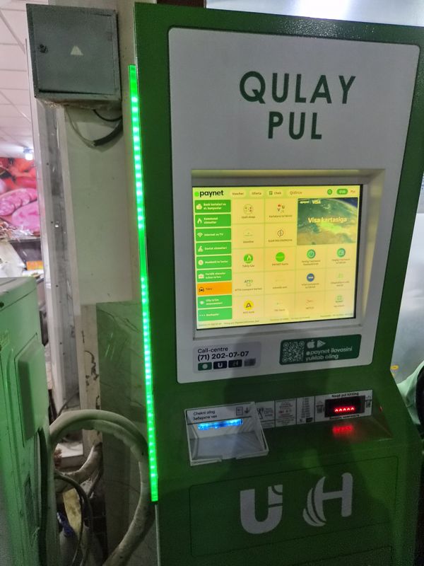 Платёжный терминал Paynet «Qulay Pul» с зелёной светодиодной полосой сбоку, интерфейс на узбекском: купюроприёмник, выдача чека; слева прилавок с колбасой и электрощиток