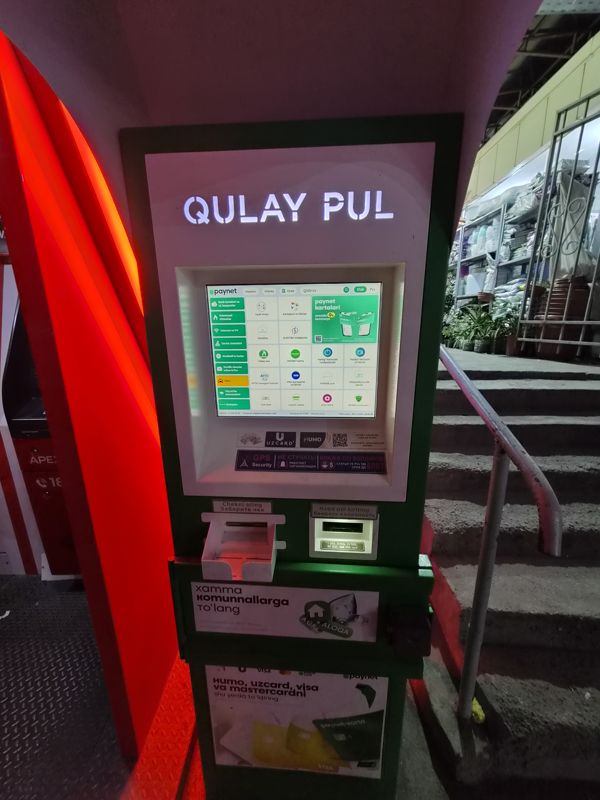 Вечером: платёжный терминал Paynet «Qulay Pul» с подсветкой надписи под навесом у лестницы базара; слева красная будка Apexbank