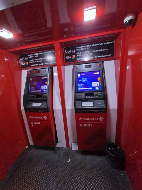 Красная будка Apexbank с двумя банкоматами рядом: над каждым табличка «обмен валют, пополнение карты, выдача наличных», купюроприёмники, видеокамера, мусорная корзина