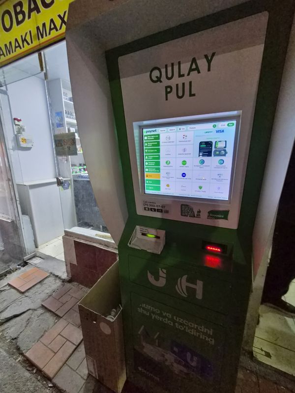 Вечером: платёжный терминал Paynet «Qulay Pul» у входа в магазин на базаре: экран с услугами, выдача чека, купюроприёмник; сверху вывеска магазина