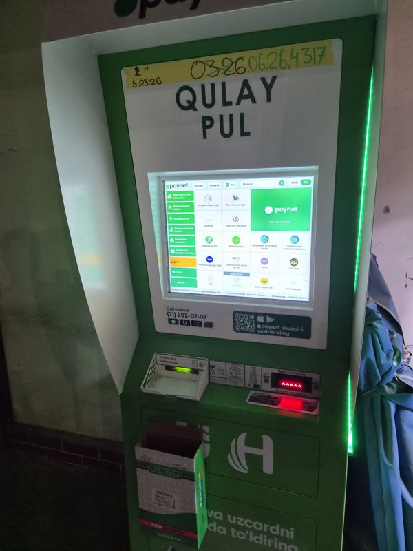 Платёжный терминал Paynet «Qulay Pul» в тёмном проходе у стены, рядом сложенный синий тент: сенсорный экран с услугами (сотовые операторы, пополнение карт, коммунальные услуги, такси), купюроприёмник, выдача квитанции, call-centre (71) 202-07-07