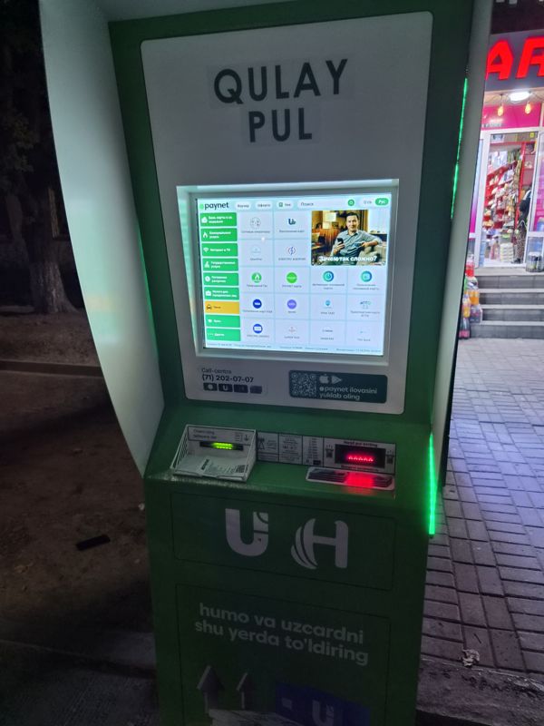 Вечером: уличный платёжный терминал Paynet «Qulay Pul» на тротуаре у ступеней входа в магазин: сенсорный экран с услугами и рекламным роликом, выдача квитанции, купюроприёмник; на корпусе «humo va uzcardni shu yerda to'ldiring», call-centre (71) 202-07-07