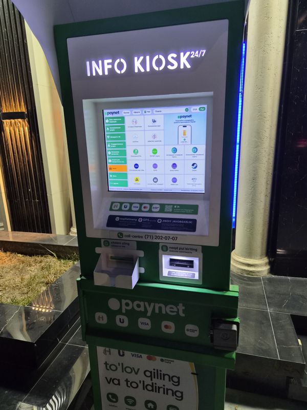 Вечером: платёжный терминал Paynet «Info Kiosk 24/7» под аркой у входа в здание с тёмной мраморной отделкой и колонной: сенсорный экран с услугами (сотовые операторы, пополнение карт, коммунальные услуги, интернет и ТВ, такси), купюроприёмник и лоток для квитанции, call-centre (71) 202-07-07, синяя подсветка сбоку