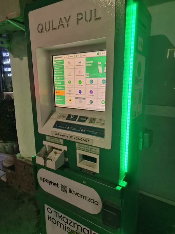 Вечером: платёжный терминал Paynet «Qulay Pul» у стены здания у стеклянной витрины, с зелёной светодиодной подсветкой сбоку: сенсорный экран с услугами, лоток квитанции, купюроприёмник (1 000–200 000 сум), наклейки Uzcard и Humo, «Paynet ilovamizda», call-centre (71) 202-07-07; слева в стекле отражаются неоновые вывески