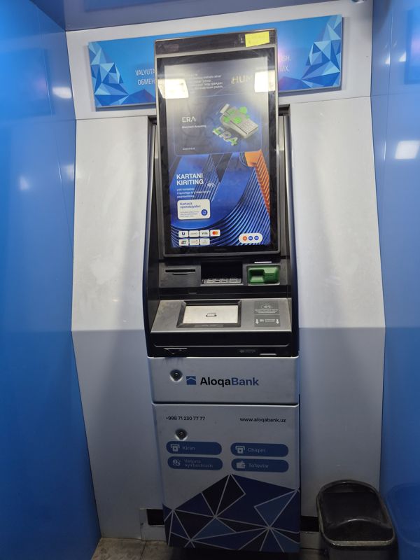 Банкомат AloqaBank (Hyosung) в синей кабине: вертикальный сенсорный экран с заставкой «Kartani kiriting» и значком бесконтактной оплаты, считыватель, купюроприёмник; на корпусе кнопки «Kirim», «Chiqim», «Valyuta ayirboshlash», «To'lovlar», www.aloqabank.uz; рядом мусорная корзина