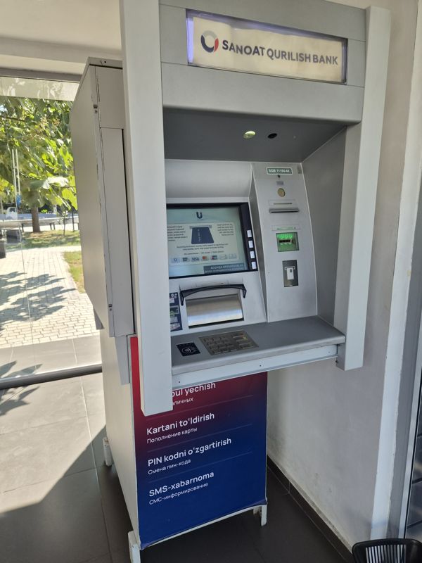 Банкомат SQB (Sanoat Qurilish Bank) в павильоне: над ним световой короб «Sanoat Qurilish Bank», на экране Uzcard инструкция вставить карту, лоток выдачи, клавиатура со знаком бесконтактной оплаты; на стойке сбоку перечень услуг: «Naqd pul yechish / Выдача наличных», «Kartani to'ldirish / Пополнение карты», «PIN kodni o'zgartirish / Смена пин-кода», «SMS-xabarnoma / СМС-информирование»