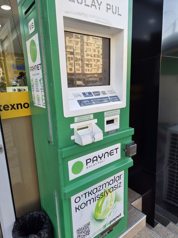 Платёжный терминал Paynet в зелёном корпусе у входа в магазин (виден жёлтый фасад «texno…»): белая панель «QULAY PUL» с экраном, логотипы Uzcard и Humo, QR приложения Paynet, наклейки «GPS Security», «Не стучать», приёмник купюр «Naqd pul kiriting» (1 000–100 000 сум), лоток чека; слева наклейка Paynet со значками услуг и метка «MS», снизу баннер «O'tkazmalar komissiyasiz», урна