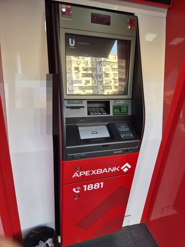Банкомат Apexbank в красной будке: большой экран Uzcard «Terminal № 113943» (на экране отражается жилой дом), клавиатура, зелёный картоприёмник, лоток купюр, знаки бесконтактных карт и штрихкода, на цоколе логотип APEXBANK и телефон 1881, сверху метка «Bankomat raqami 091», у стены урна