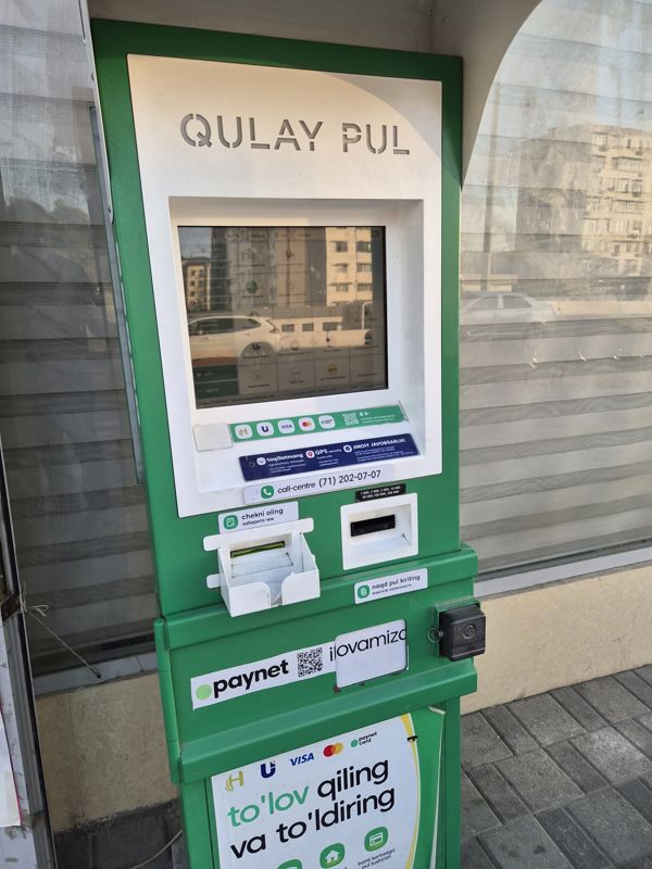 Платёжный терминал Paynet «Qulay Pul» в зелёно-белом корпусе у стеклянной стены павильона: сенсорный экран с услугами (сотовые операторы, пополнение карт, электроэнергия, Uzum, такси и др.), приём наличных, выдача чека, наклейки Paynet и QR, Call-centre (71) 202-07-07, логотипы Humo, Uzcard, Visa, Mastercard, Paynet Card; на экране отражение улицы