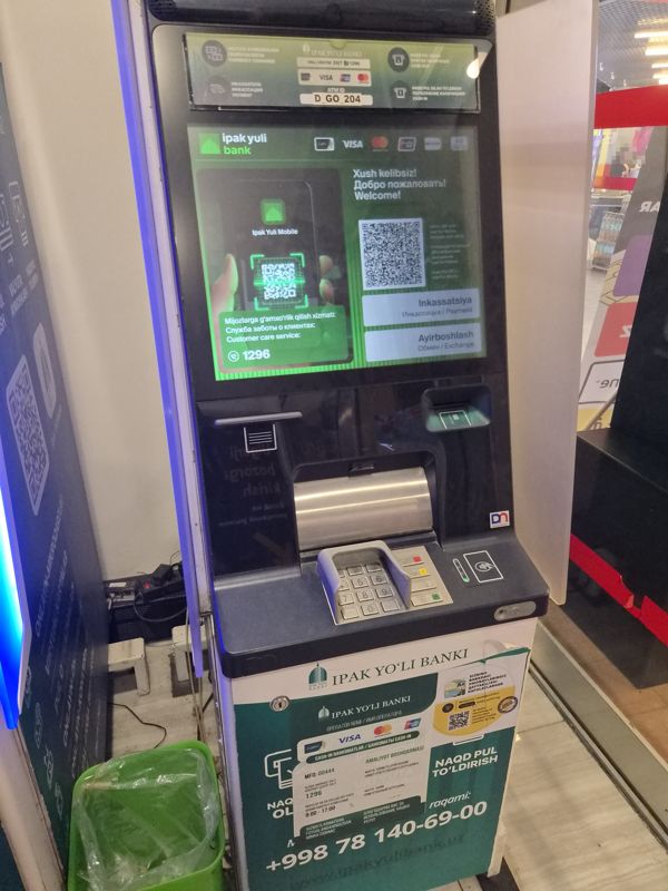 Внутри ТРЦ «Сампи Молл»: банкомат Ipak Yuli Bank с зелёным экраном «Xush kelibsiz! / Добро пожаловать!», QR-кодом приложения Ipak Yuli Mobile и кнопками «Инкассация» и «Обмен»; над экраном ATM ID D_GO_204, ниже клавиатура с бесконтактным считывателем, на корпусе наклейки с телефонами и режимом работы; справа у входа в Корзинку видна касса магазина