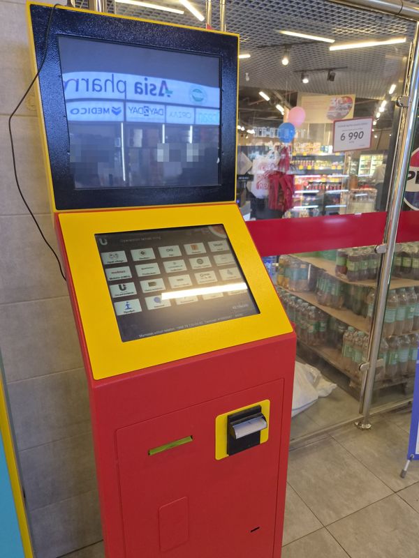 Внутри ТРЦ «Сампи Молл»: жёлто-красный платёжный терминал с двумя экранами у стеклянного входа в Корзинку; на сенсорном экране выбор оператора (Ucell, Beeline, Humans, UzMobile, Mobiuz, Perfectum, SQB, Uzcard, Humo, Visa, коммунальные платежи, интернет, бюджет), внизу телефон +998 78 120-00-82 и номер терминала; ниже щель приёма купюр и принтер чеков; на верхнем экране отражаются вывеска Asia pharm и аптечные витрины
