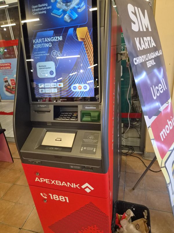 Внутри ТРЦ «Сампи Молл»: банкомат Apexbank в красном корпусе с большим экраном «Kartangizni kiriting» (Uzcard, Humo, Visa, Mastercard, UnionPay), под ним клавиатура, зелёный картоприёмник, лоток купюр, знаки бесконтактных карт и штрихкода, на цоколе телефон 1881; справа баннер Ucell «SIM karta — chiroyli raqamlar bepul», у стены урна