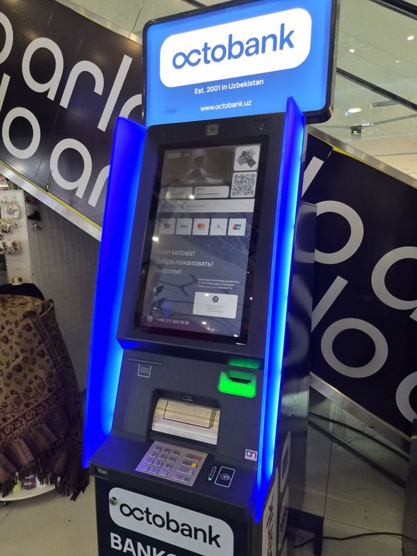 Внутри ТРЦ «Сампи Молл» у эскалатора: банкомат Octobank с синей подсветкой и светящейся вывеской «octobank — Est. 2001 in Uzbekistan, www.octobank.uz»; на экране «Xush kelibsiz! / Добро пожаловать!», логотипы Visa, Mastercard, Uzcard, UnionPay, QR и телефон +998 (71) 202 33 33; ниже лоток купюр, клавиатура и бесконтактный считыватель; фоном эскалатор с чёрными вывесками