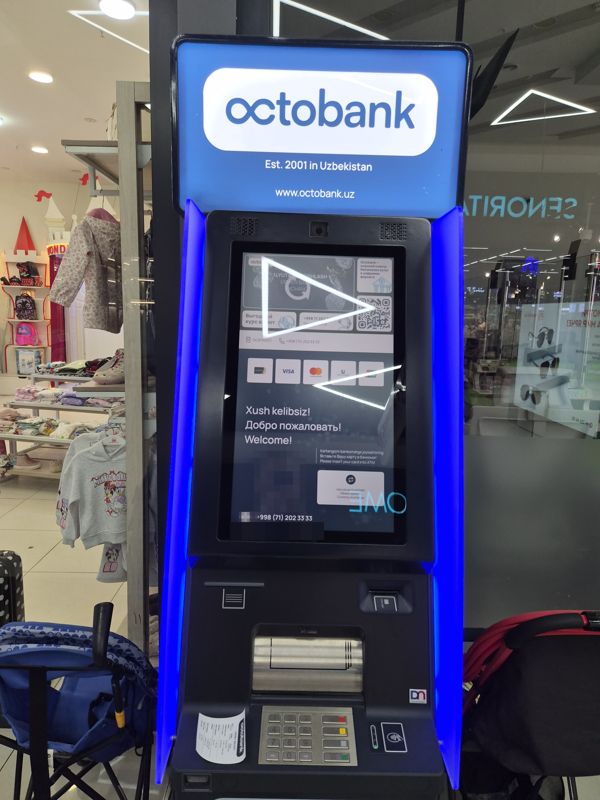 Внутри Экобазара: банкомат Octobank с синей подсветкой и светящейся вывеской «octobank — Est. 2001 in Uzbekistan, www.octobank.uz»; на экране «Xush kelibsiz! / Добро пожаловать! / Welcome!», реклама обмена валют по выгодному курсу, QR, логотипы Humo, Visa, Mastercard, Uzcard, UnionPay, кнопка «Обмен валют», номер OCBTM031 и телефон +998 (71) 202 33 33; ниже картоприёмник, лоток купюр, клавиатура и бесконтактный считыватель; слева магазин детской одежды, справа витрина «Senorita»