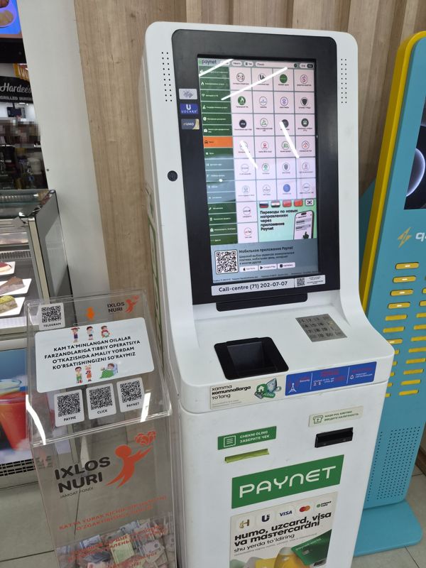 Внутри Экобазара у фудкорта (видна вывеска Hardee's): белый платёжный терминал Paynet с сенсорным экраном услуг (сотовые операторы, коммунальные услуги, интернет, штрафы, такси, кредиты и др.), приём наличных, принтер чеков; наклейки «Humo, Uzcard, Visa va Mastercardni shu yerda to'ldiring», Call-centre (71) 202-07-07, на экране «Терминал: 9138570»; слева прозрачный ящик для пожертвований фонда «Ixlos Nuri», справа стойка q.watt