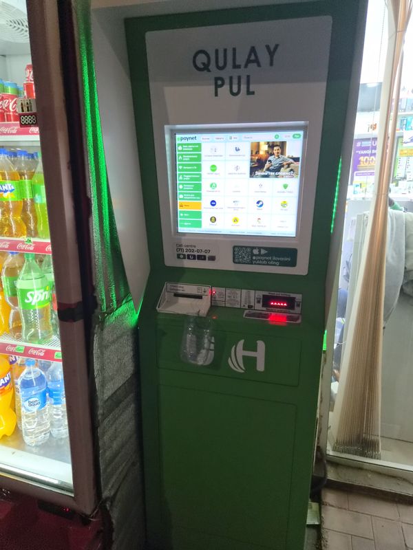Платёжный терминал Paynet с надписью «Qulay pul» в магазине у холодильников с напитками; экран с меню оплаты услуг, приём наличных, печать чека. Наличные не выдаёт.