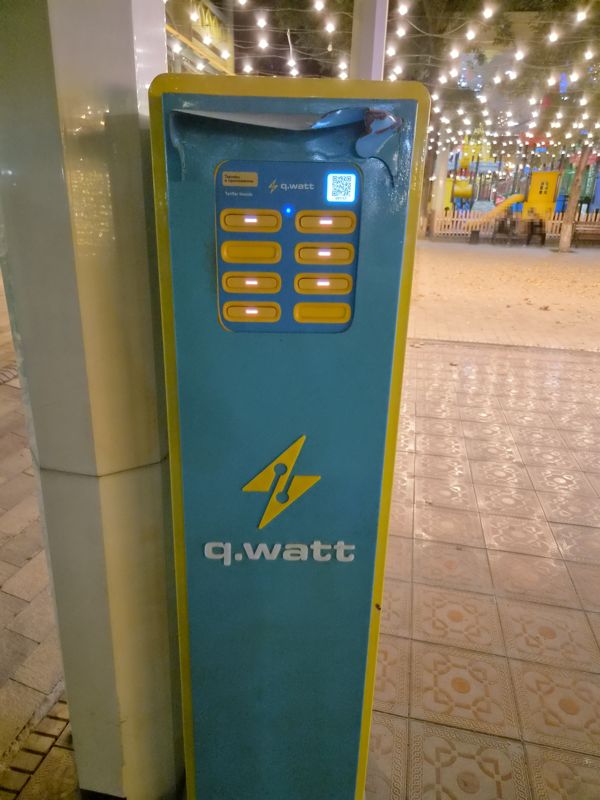 Стойка проката повербанков q.watt с восемью ячейками и QR-кодом у столба возле детской площадки; тарифы в приложении («Tariflar ilovada»)