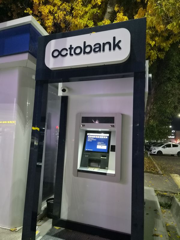 Вечером: банкомат Octobank в уличной будке с тёмно-синей рамой и светящейся вывеской «octobank»; на экране «Xush kelibsiz! / Добро пожаловать! / Welcome!» и логотипы Visa, Mastercard, Uzcard, UnionPay, ниже картоприёмник, лоток купюр и клавиатура; над банкоматом камера наблюдения