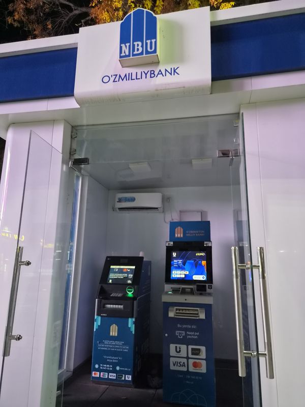 Два банкомата NBU (O'zmilliybank) в стеклянной уличной будке с вывеской «NBU O'zmilliybank»: слева банкомат с выдачей наличных в сумах по картам, справа банкомат с экраном Uzcard / Humo и приёмом наличных
