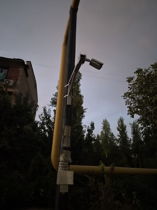 Столб с жёлтой газовой трубой во дворе: цилиндрическая камера на кронштейне, объявления внизу