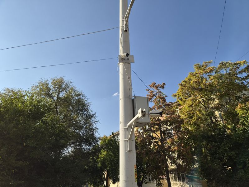 Днём: тот же столб с другого ракурса: на кронштейне у шкафа цилиндрическая камера, выше на хомуте маленькая камера; вокруг деревья и жилой дом