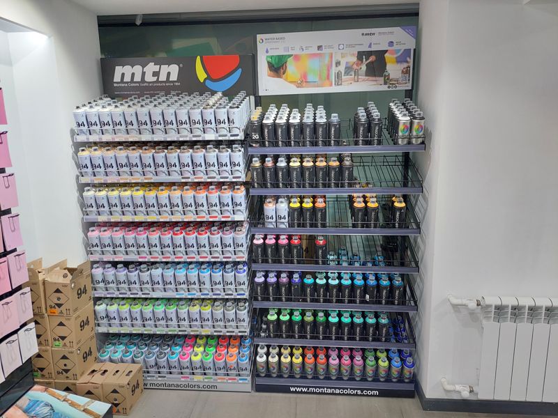 Стенд аэрозольных красок mtn (Montana Colors): слева ряды баллонов серии 94 по цветам, справа стеллаж с Hardcore, Nitro 2G и Water Based; над ними вывеска mtn и плакат Water Based Spraypaint 400, внизу «www.montanacolors.com»