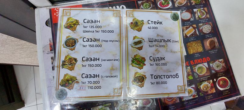 Меню ресторана на столе: рыбные блюда по килограммам (сазан, судак, толстолоб), стейк и шашлык из сома; за ним фотоменю закусок, лепёшек и супов