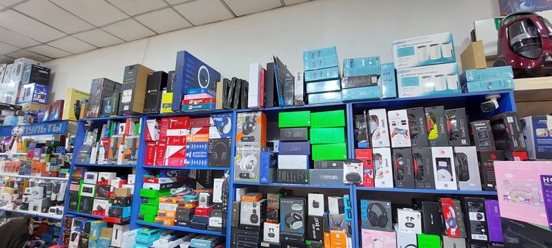 Стены магазина электроники: полки с игровыми наушниками, мышами, клавиатурами, микрофонами; сверху роутеры TP-Link, кольцевая лампа, коробки PS5