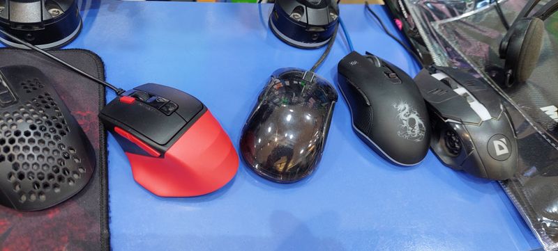 Игровые мыши на столе: красно-чёрная, прозрачная, две Defender; рядом гарнитура и коврик в упаковке