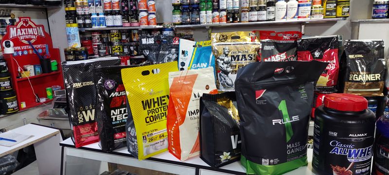 Прилавок с пакетами протеина и гейнеров; на стене вывеска Maximum Nutrition (vistansport.uz), за прилавком полки с витаминами