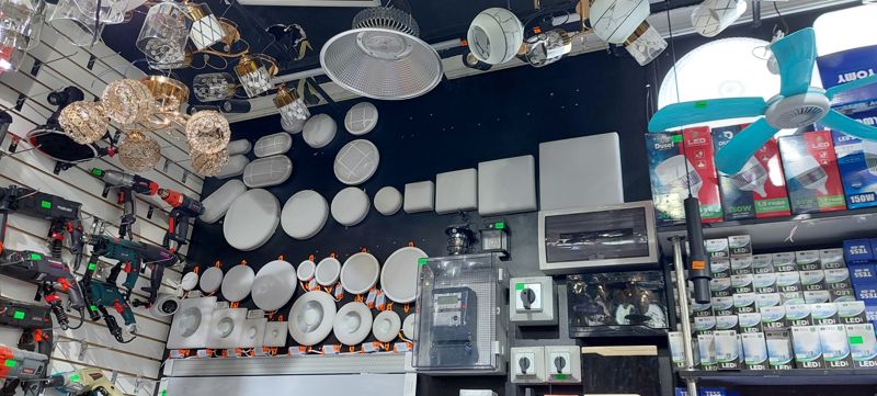 Стена светильников: накладные круглые, овальные и квадратные LED-светильники, точечные COB, люстры; электрощиток с электросчётчиком в шкафу, пакетные переключатели, потолочный мини-вентилятор бирюзовый, лампы Dusel и Tess; слева дрели и перфораторы с ценниками