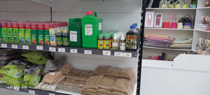 Полка с жидкими удобрениями Bona Forte, Зелёное мыло и Green Belt Климат, канистра Agroimmun, флаконы Агрикола и Биогумат над рядом ценников; ниже мешки коры сосны и фасованная по пакетам крупа-подкормка; справа за стойкой флористические материалы: бумага, ленты, коробки, корзинки