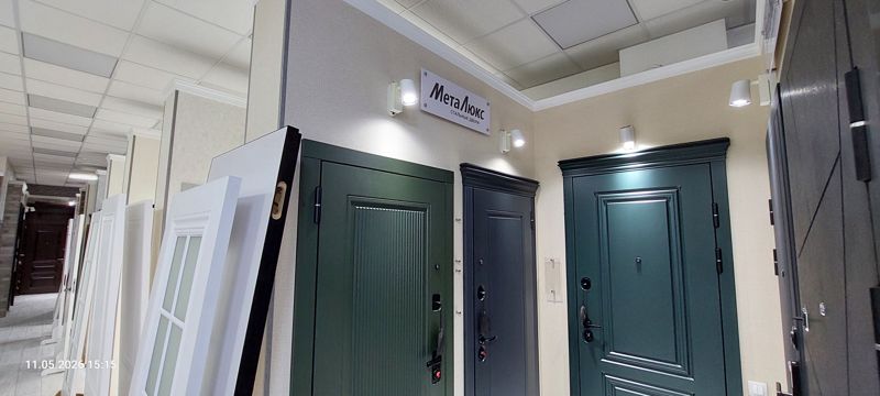 Зал с образцами стальных дверей под вывеской «МетаЛюкс — стальные двери»: зелёная ребристая, серая, тёмно-зелёная; слева белые межкомнатные двери
