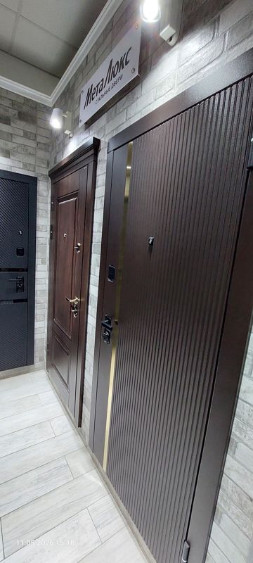 Образцы стальных входных дверей на фоне светлой кирпичной стены под вывеской «МетаЛюкс — стальные двери»: коричневая дверь с вертикальным рифлением и золотистой вставкой, дверь цвета тёмного дерева с наличником и филёнками