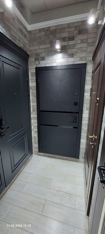 Образцы стальных входных дверей на фоне светлой кирпичной стены: по центру чёрная дверь с диагональным рифлением и горизонтальной вставкой, по бокам тёмно-серая дверь с филёнками и дверь цвета тёмного дерева