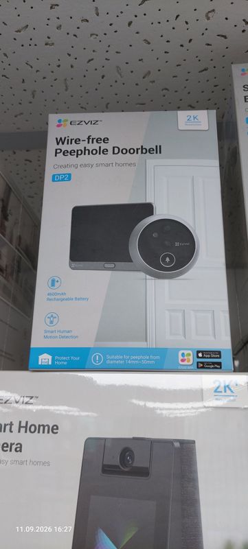 Коробка умного дверного глазка-звонка EZVIZ DP2 (Wire-free Peephole Doorbell, 2K) на полке; ниже виден край коробки умной камеры EZVIZ Smart Home Camera
